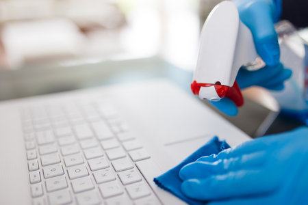 Close up of female hands in surgical protective gloves cleaning laptop keyboard with microfiber cloth and spray disinfectantの写真素材