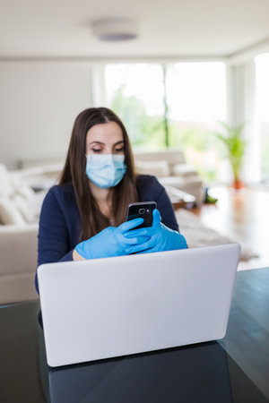 Young businesswoman with surgical mask and gloves using mobile phone and working online from home, covid-19 isolation concept.の写真素材
