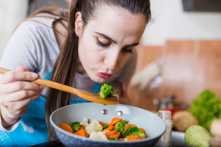 Young housewife preparing meal of mixed vegetables and tasting broccoli with mixing spoon in kitchen.の写真素材