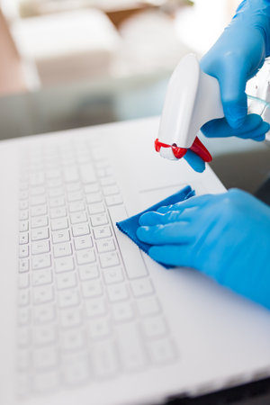 Woman wearing medical rubber gloves wiping laptop keyboard with small rag and using spray bottle sanitizer alcohol, laptop disinfection conceptの写真素材