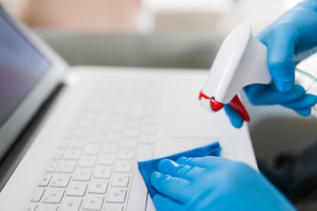 Close up of female hands in surgical protective gloves cleaning laptop keyboard with microfiber cloth and spray disinfectantの写真素材