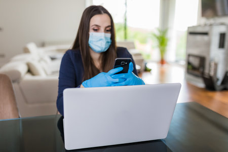Young business woman working from home on laptop and using smartphone, wearing protective mask and gloves. Coronavirus quarantine concept.の写真素材