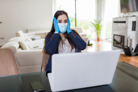 Stressed woman wearing face mask and protective gloves while working on laptop at home. Covid-19 and business problems concept.の写真素材