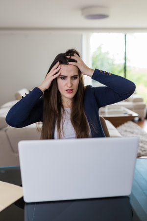 Worried young woman in front of laptop in living room. Fail and technology concept.の写真素材