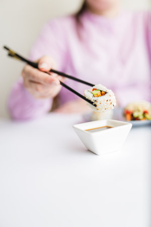 Female dipping tasty sushi roll with chopsticks into bowl with soy sauce at the table.の写真素材
