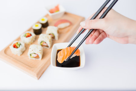 Close up of woman holding chopsticks with salmon sushi roll and bowl with soy sauce on table, portion of traditional japanese meal in the background.の写真素材