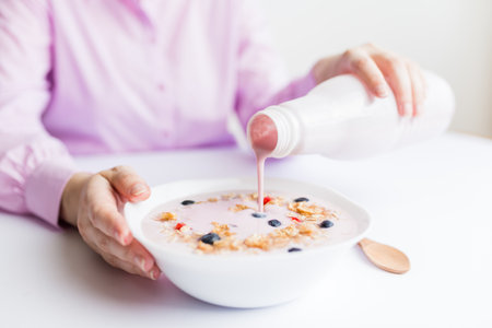 Close up of female pouring fruit yogurt into bowl with cereals, strawberries and blueberries.の写真素材
