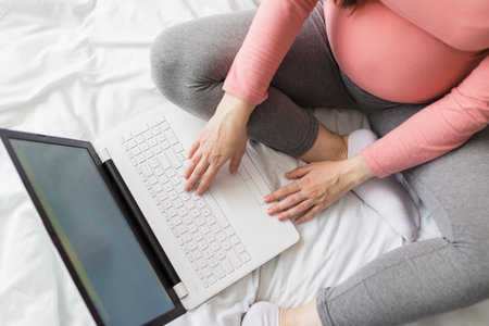 High angle view of young pregnant woman sitting on bed and using laptop.