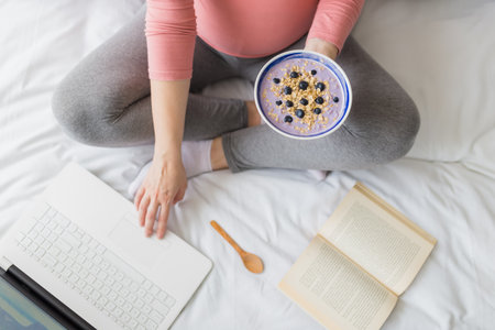 Close up of pregnant woman eating muesli with blueberries while working on laptop and reading book.の写真素材