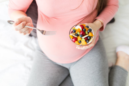 High angle view of pregnant female sitting in bedroom and eating fresh fruit salad.の写真素材
