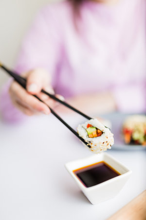 Female dipping tasty sushi roll with chopsticks into bowl with soy sauce at the table.の写真素材