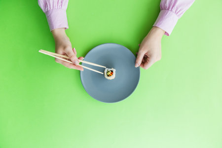Flat lay of female hand holding chopsticks and one sushi roll on plate isolated on green background.の写真素材