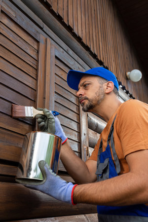 Worker applying protective varnish or wood oil on wooden house cottage exterior walls and window shuttersの写真素材