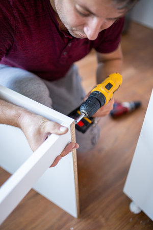 Furniture Installation. Man Using Electric Drill Assembling And Fixing Wooden Shelf Furnishing Room...