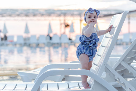 Baby girl on summer vacation, standing on lounger next to swimming pool. Sunset in the background.の写真素材