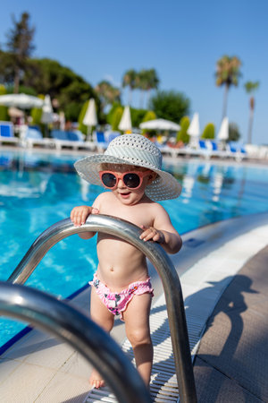 Fashionable baby girl standing near outdoor swimming pool at tourist resort. Summer vacation concept.の写真素材