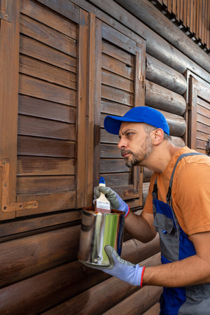 Worker applying protective varnish or wood oil on wooden house cottage exterior walls and window shuttersの写真素材