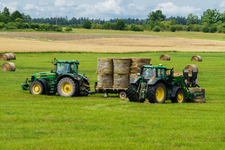 A tractor loads bales of hay onto a trailer from another tractor. Kurzeme,Latvia, July 15, 2019.のeditorial素材
