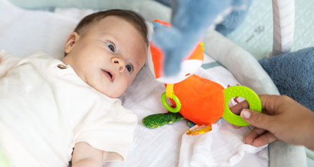 Adorable little baby lying on children rug and looking at toy hold by his mother hand