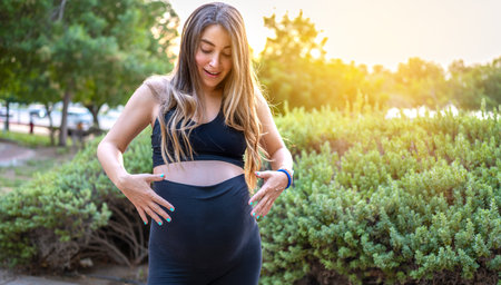 Excited future mother looking at her belly with arms around while standing at park.の写真素材