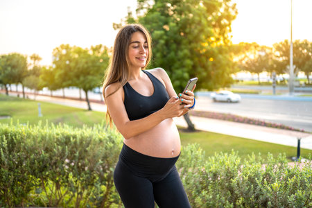 Portrait of smiling pregnant woman in sportswear using phone outdoors at sunset.