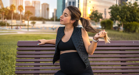 Portrait of pregnant woman resting on wooden park bench at sunset.