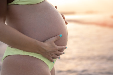 Close up of pregnant woman belly wearing yellow bikini on the beach at sunset.の写真素材