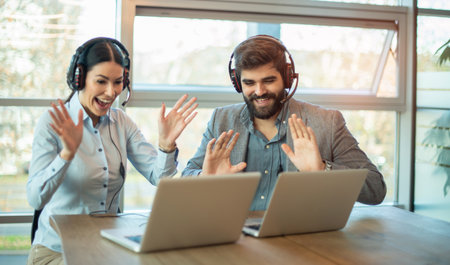 Coworkers smiling and waving hands while talking to friend on laptop during online virtual meetingの写真素材