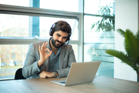 Multicultural businessman wearing call center agent headset showing OK sign with fingers to laptops screen.の写真素材
