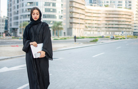 Beautiful young Arabian woman holding laptop and phone on the street.の写真素材