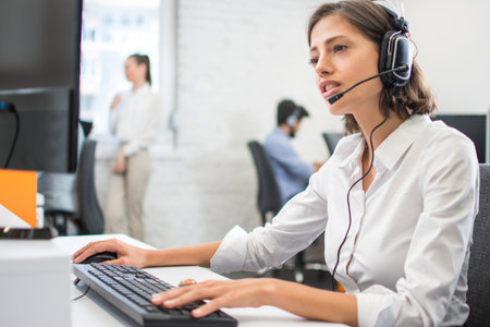 Beautiful friendly female customer support operator with headset using computer in call center.の写真素材