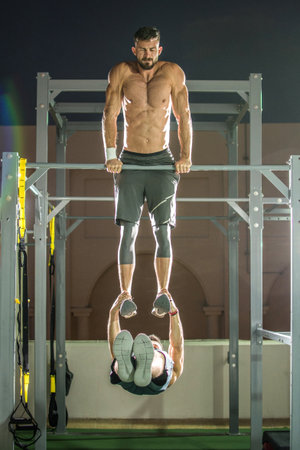 Strength and endurance exercises, man on top doing dips on horizontal bar while male friend hanging...