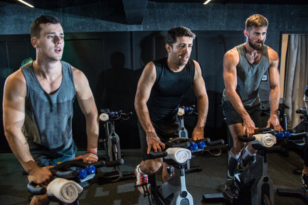 Three athletic men exercising on bikes at gym.