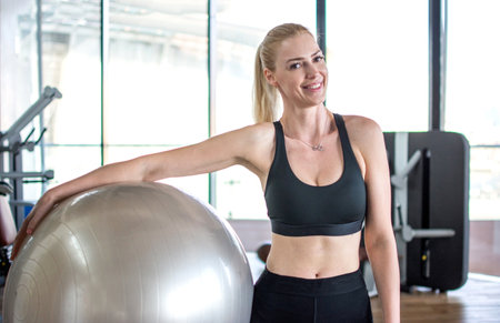 Sporty blonde woman holding fitness ball at gym. Young smiling girl standing and holding pilates ball in gym.の写真素材