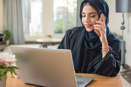 Beautiful young middle eastern arab woman talking on mobile phone and using laptop at home.