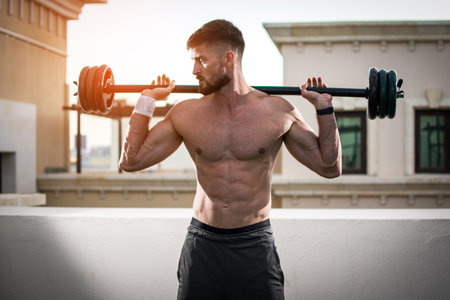 Muscular sexy man with barbell over shoulders working out outdoors