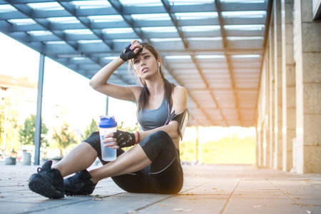 Tired sporty girl with water bottle sitting down on sidewalk after workout in urban area.