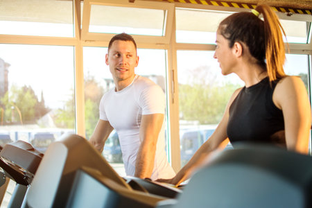 Sporty friends walking on treadmill and talking at gym.