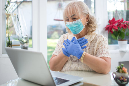 Scared mid age woman wearing protective mask and gloves reading news on laptop at home.の写真素材