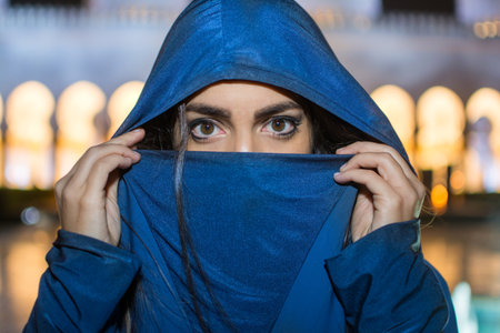 Headshot of beautiful Muslim woman in traditional Arab clothes covering her face near Mosque during...
