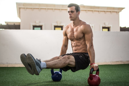 Muscular shirtless man workout with kettlebells in L Sit position outdoors