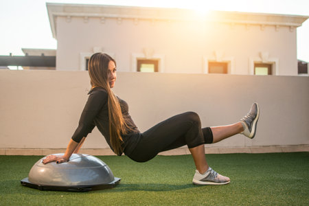 Long haired slim fit sportswoman exercising on bosu ball outdoors during sunset