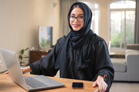 Middle eastern woman wearing abaya with laptop and smart phone looking at camera while sitting near...
