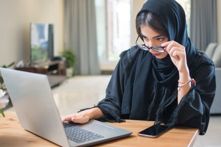 Pretty young middle eastern woman wearing abaya and eyeglasses using laptop at home.の写真素材