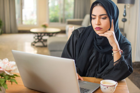 Bored young arab woman wearing hijab with a laptop staring at screen with concerned facial expression indoorsの写真素材