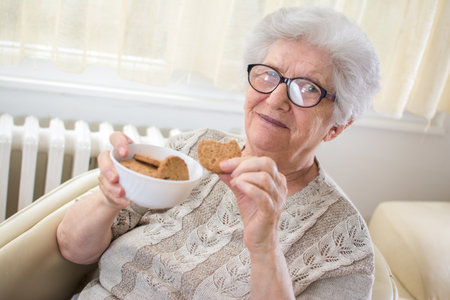 Portrait of smiling senior woman holding a plate with cookies and enjoying delicious biscuit at homeの写真素材