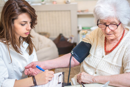 Nurse measuring blood pressure of senior woman at home. Female doctor writing medical results to...