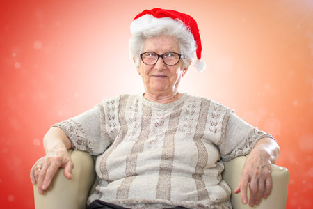 Happy senior woman with red Santas hat sitting in sofa against red Xmas background.の写真素材