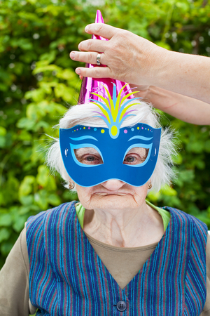 Portrait of a happy elderly woman celebrating birthday outdoor, wearing a colorful hat and maskの写真素材