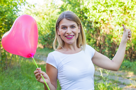 Picture of a smiling beautiful woman holding a red helium balloon ans showing thumbs up outdoorの写真素材